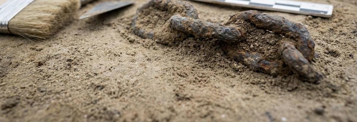 Detalle macro de una cadena de hierro antigua junto a sedimento y herramientas de registro arqueológico, con foco en textura y uso