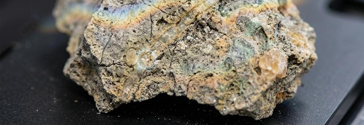 Macro editorial de una muestra rocosa con vetas minerales y una lectura espectral sutil, evocando una posible alteración por agua sin afirmarla.