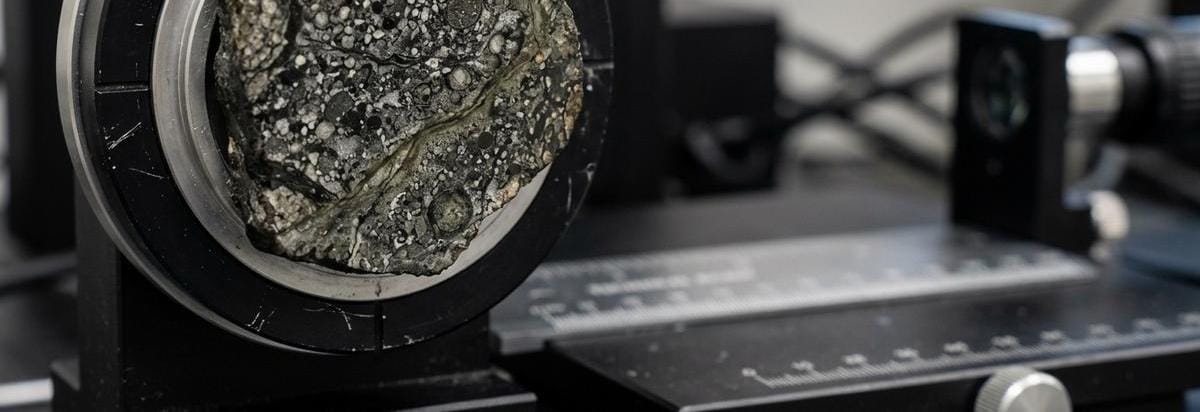 Sección pulida de un meteorito con vetas minerales y pequeñas inclusiones, iluminada para sugerir señales de agua antigua.