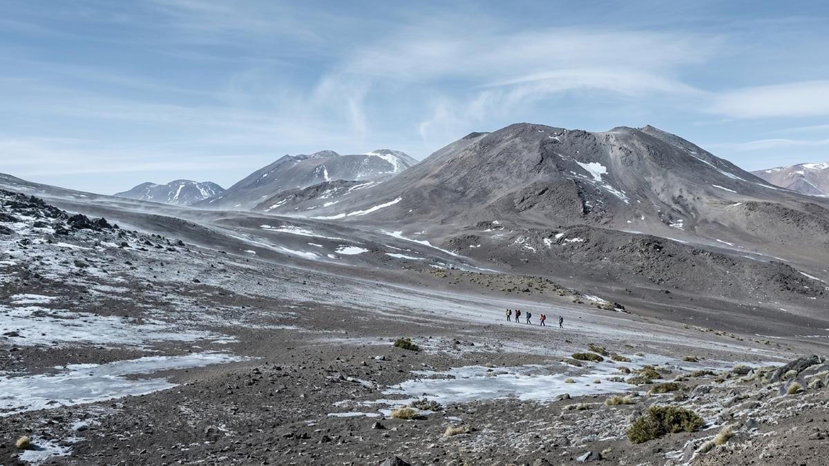 Paisaje volcánico helado de la Puna de Atacama con una pequeña expedición científica a distancia, mostrando escala y aislamiento.