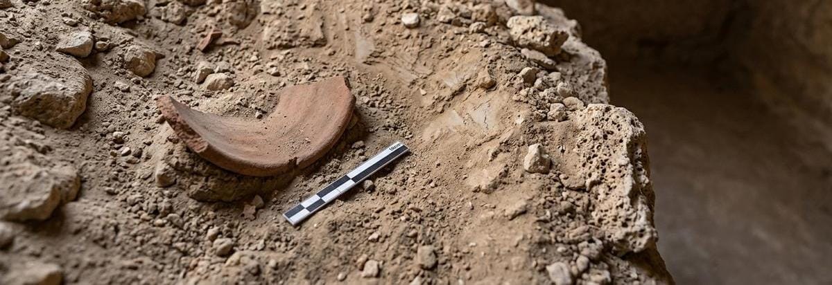 Detalle arqueológico de una cámara funeraria con estratos de roca, marcas de excavación y un fragmento de cerámica junto a una regla de escala