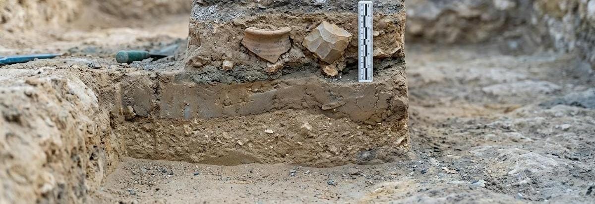 Detalle arqueológico de una tumba parcialmente excavada con capas de sedimento, perfil estratigráfico y una herramienta de medición discreta junto a fragmentos de cerámica.