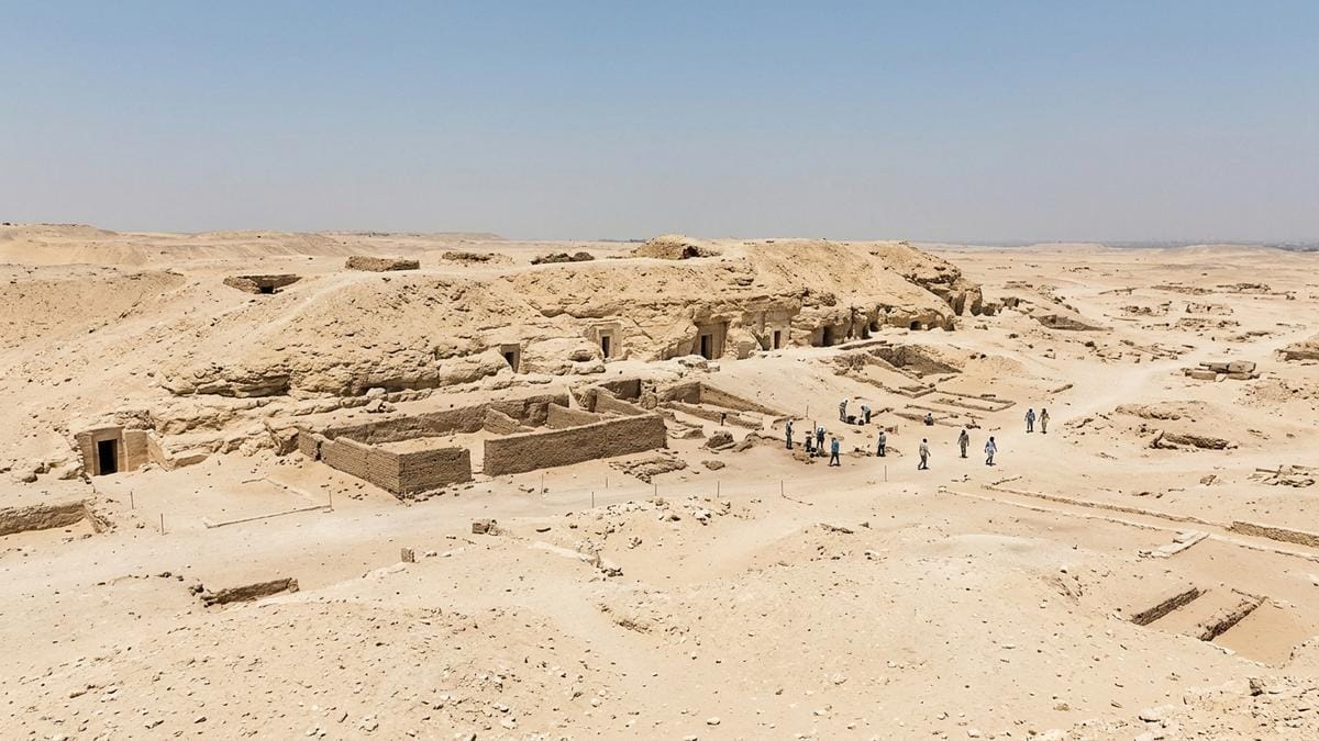 Panorámica de la necrópolis de Saqqara con una ladera de roca caliza, restos de tumbas excavadas y arqueólogos trabajando a distancia bajo luz diurna suave.