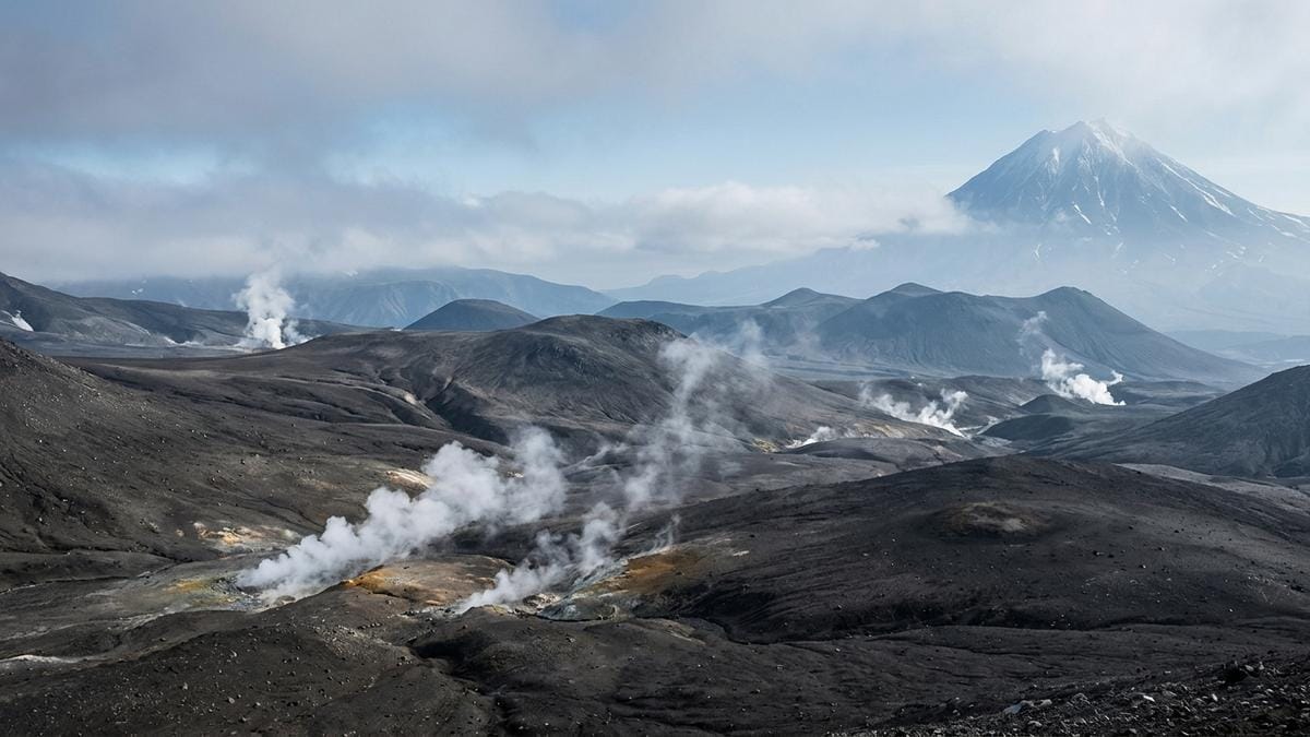 Panorámica del paisaje volcánico de Kamchatka con vapor bajo y laderas oscuras, sin protagonismo de microhongos, como contexto geológico del estudio en el Avachinsky.